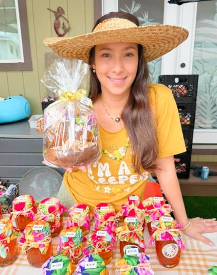 Kristal in a straw hat at the market table, holding a gift basket above rows of fabric-topped honey jars